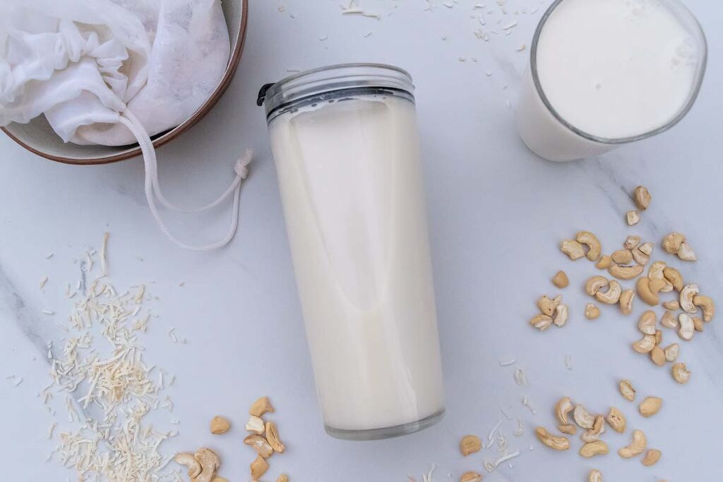 Cashews and coconut scattered around milk made in thermomix, in a jar laying on its side and a glass. Off to the top left is a bowl containing a nut milk bag and the pulp from making the milk