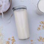 Cashews and coconut scattered around milk made in thermomix, in a jar laying on its side and a glass. Off to the top left is a bowl containing a nut milk bag and the pulp from making the milk