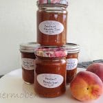 four jars thermomix peach jam on a white plate with white background. Peaches beside them.