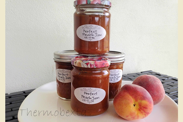 four jars thermomix peach jam on a white plate with white background. Peaches beside them.