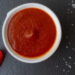 White bowl containing red sauce, sliced tomato and rock salt all sitting on black textured tile