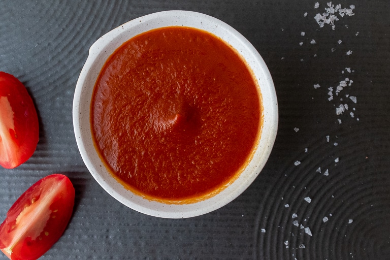 White bowl containing red sauce, sliced tomato and rock salt all sitting on black textured tile