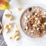 thermomix overnight oats in glass bowl with blueberries on top. Sitting on white round board on top of rustic black table. Pear, blueberries and cashews around