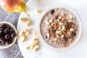 thermomix overnight oats in glass bowl with blueberries on top. Sitting on white round board on top of rustic black table. Pear, blueberries and cashews around