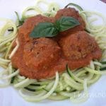 Four meatballs on top of zoodles, topped with tomato sauce and two basil leaves. On white plate.