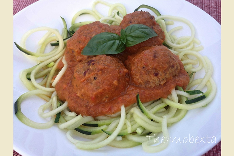 Four meatballs on top of zoodles, topped with tomato sauce and two basil leaves. On white plate.