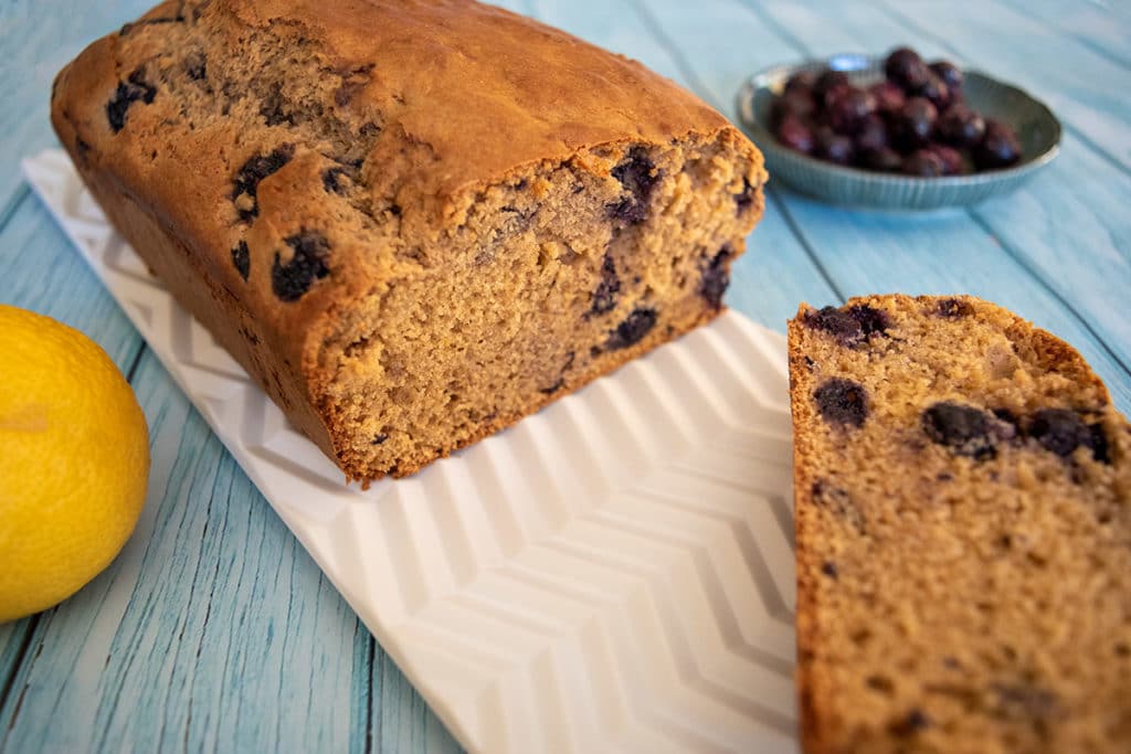 blueberry and lemon loaf on white platter on aqua tabletop, with one slice cut off. Lemon and small bowl of blueberries nearby