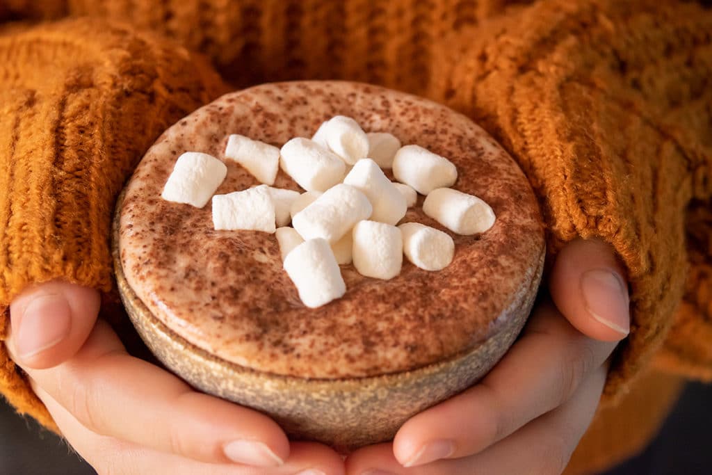 thermomix hot chocolate in mug with marshmallows being held with two hands, person wearing snuggly orange jumper