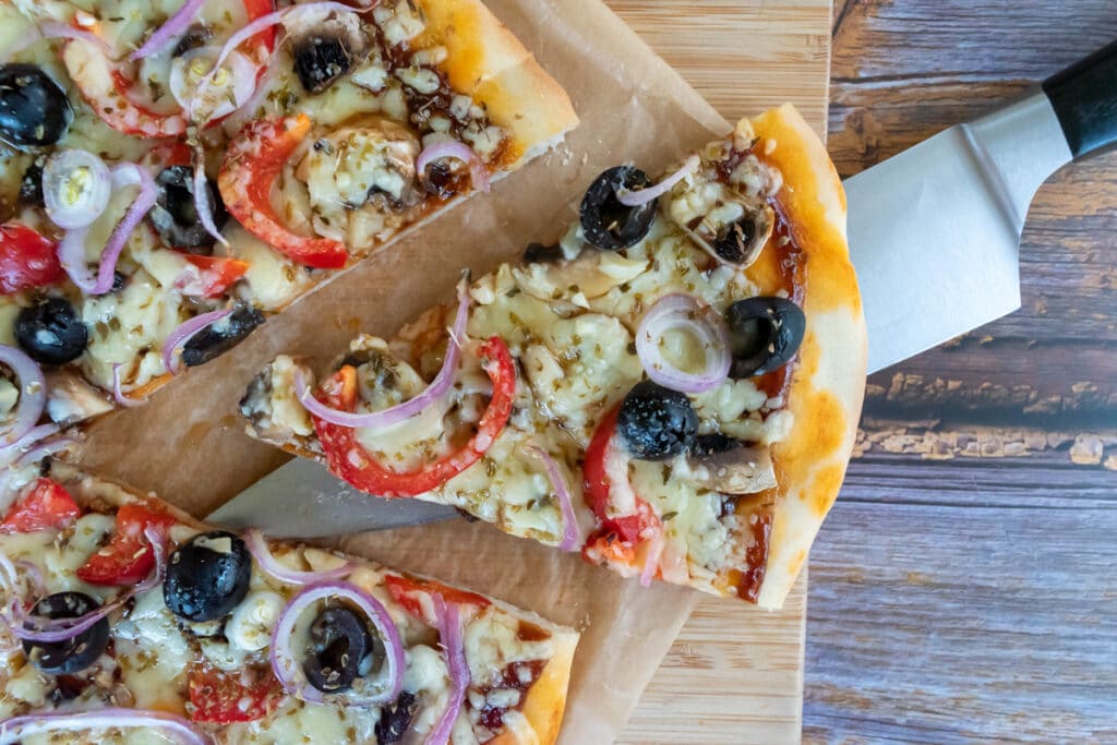 looking down on pizza that is on top of parchment paper on a light coloured wooden board on a darker wooden tabletop. Pizza has a slice cut out and that slice is sitting on top of a blade of a large knife