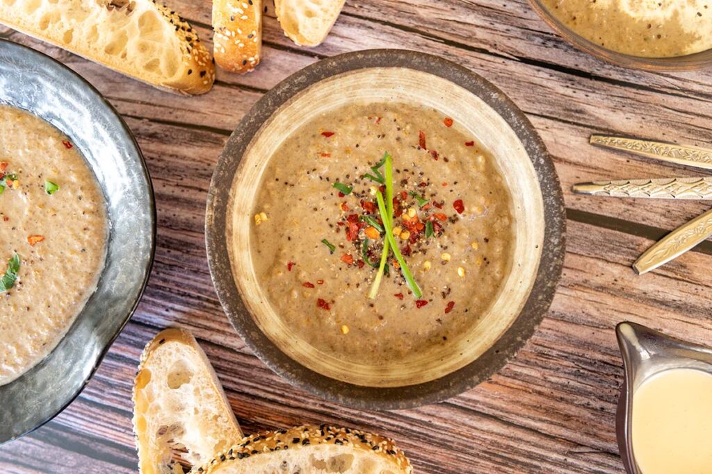 top down photo of a brown bowl of mushroom soup, topped with chilli flakes and chives, parts of two other bowls off to the sides and pieces of turkish bread