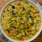 Birdseye view of thermomix vegetable casserole showing a creamy casserole in a white dish, topped with lots of finely chopped parsley. Sitting on a tea towel on a wooden board