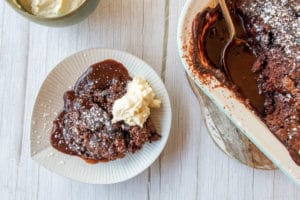 chocolate self saucing pudding portion on white plate with cream. Dish of pudding and bowl of cream off to the sides.