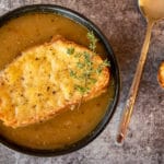 french onion soup in black bowl, with cheesie on top, spoon on right with cheesie right of the spoon