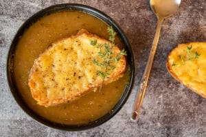 french onion soup in black bowl, with cheesie on top, spoon on right with cheesie right of the spoon