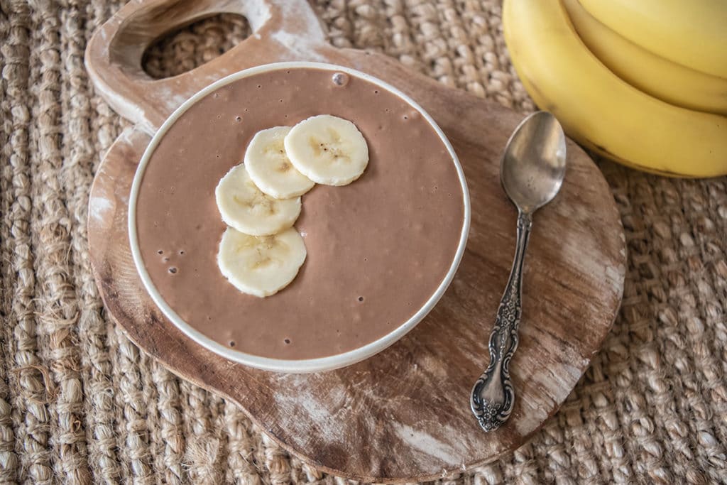 choc banana custard in bowl with four slices of banana on top, sitting on rustic wooden board on woven placemat, two bananas off to the side