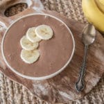 choc banana custard in bowl with four slices of banana on top, sitting on rustic wooden board on woven placemat, two bananas off to the side