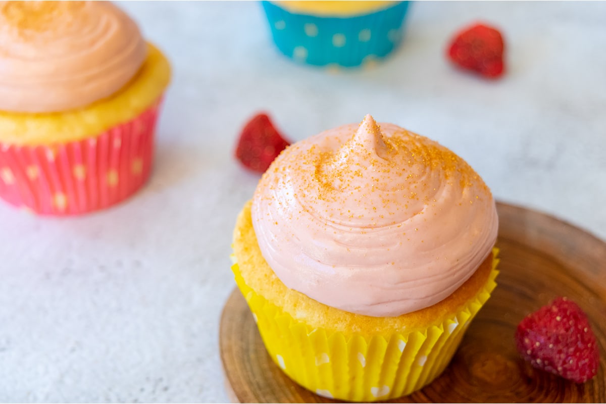 strawberry lime cream cheese frosting on cupcake in yellow case, with gold dust on top. Sitting on small round wooden plate on light table with parts of two cupcakes and two freeze-dried strawberries in the background