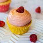 cream cheese iced cupcake with freeze-dried strawberries around, one on top of the cupcake. Cake is sitting on a detailed white platter and there is part of another cupcake in the background