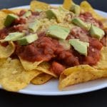 white plate containing nachos - corn chips, red sauce, avocado and grated cheese