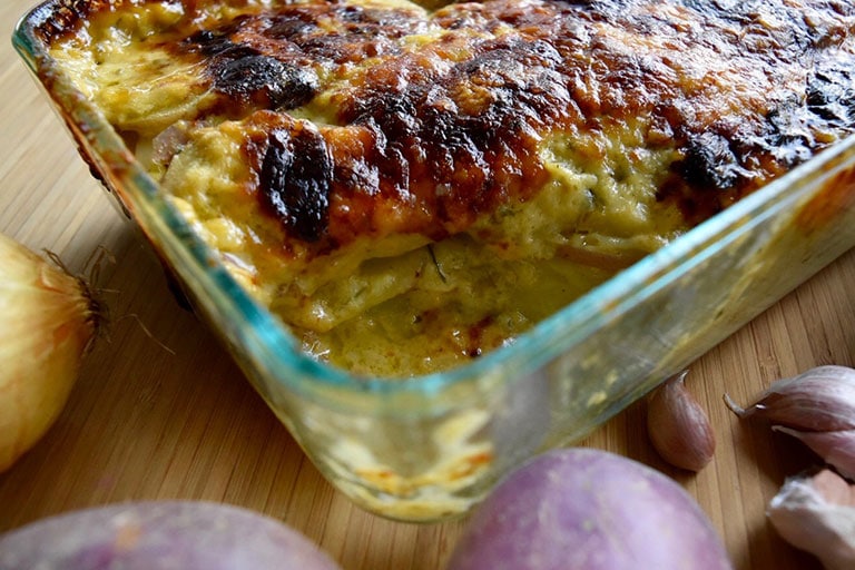 thermomix potato bake in clear dish with corner taken out of the bake. Sitting on wooden board, with part of potatoes and onions off to the side