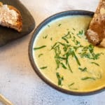 thermomix broccoli soup in black bowl with toast in it and herbs on top, toast in background on black plate. grey background