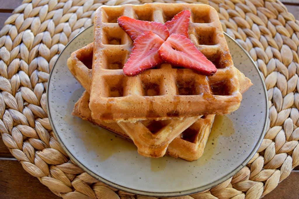 three thermomix waffles on grey plate on straw woven round placemat. Maple syrup and strawberries on top of waffles