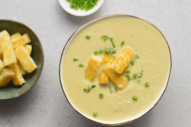 thermomix cauliflower soup in bowl topped with halloumi and chives. White background. Bowl of halloumi and bowl of chives next to soup.