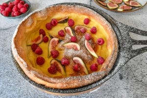 dutch baby pancake in black cast iron pan on rustic background. Fruit inside and outside on plates