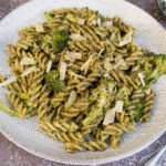 Pesto pasta and broccoli on white plate with grey detail. Rustic dark background, small bowl of cheese in background