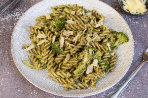 Pesto pasta and broccoli on white plate with grey detail. Rustic dark background, small bowl of cheese in background