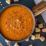 spicy tomato and lentil soup made in thermomix, in brown bowl with handle. Sitting on black slate tile on grey backdrop, with croutons and dry lentils in on the tile