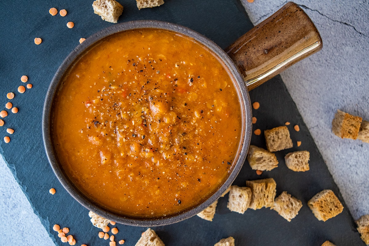 spicy tomato and lentil soup made in thermomix, in brown bowl with handle. Sitting on black slate tile on grey backdrop, with croutons and dry lentils in on the tile