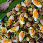 Salad of spinach, potatoes, avocado, boiled eggs, olives, on blue platter sitting on green leaf placemat on white tabletop, above platter are a set of salad servers and a jar containing yellow and green dressing