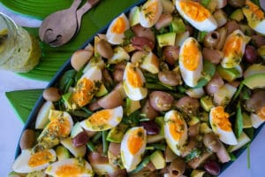 Salad of spinach, potatoes, avocado, boiled eggs, olives, on blue platter sitting on green leaf placemat on white tabletop, above platter are a set of salad servers and a jar containing yellow and green dressing