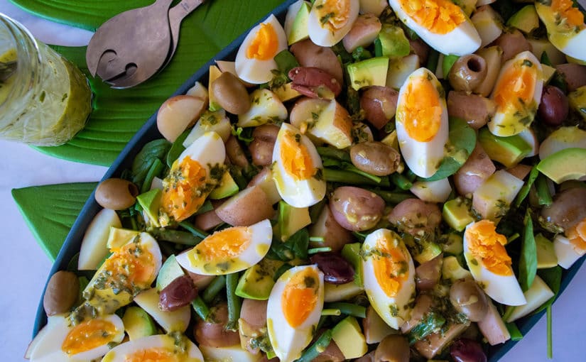 Salad of spinach, potatoes, avocado, boiled eggs, olives, on blue platter sitting on green leaf placemat on white tabletop, above platter are a set of salad servers and a jar containing yellow and green dressing