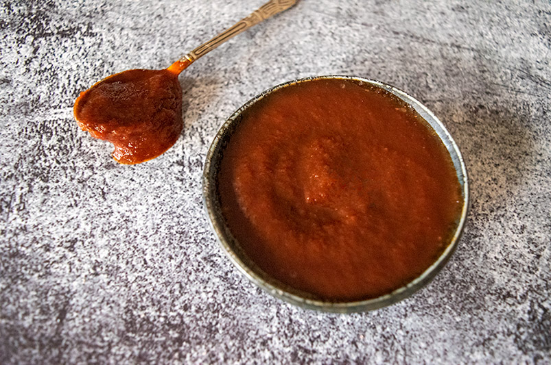 rustic grey/black backdrop, sitting on top is faded black ramekin with Thermomix bbq sauce in it, spoon on surface as well, off to the side top left with bbq sauce on it, spilling onto table