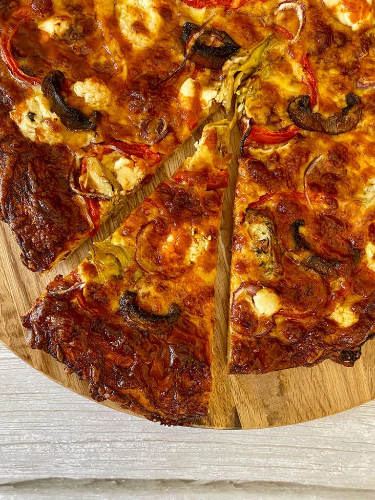 bottom left quarter of pizza on wooden round board on grey wooden countertop. Pizza is topped with Thermomix bbq sauce, mushrooms, artichokes, red onion, red capsicum, Persian feta and mozzarella