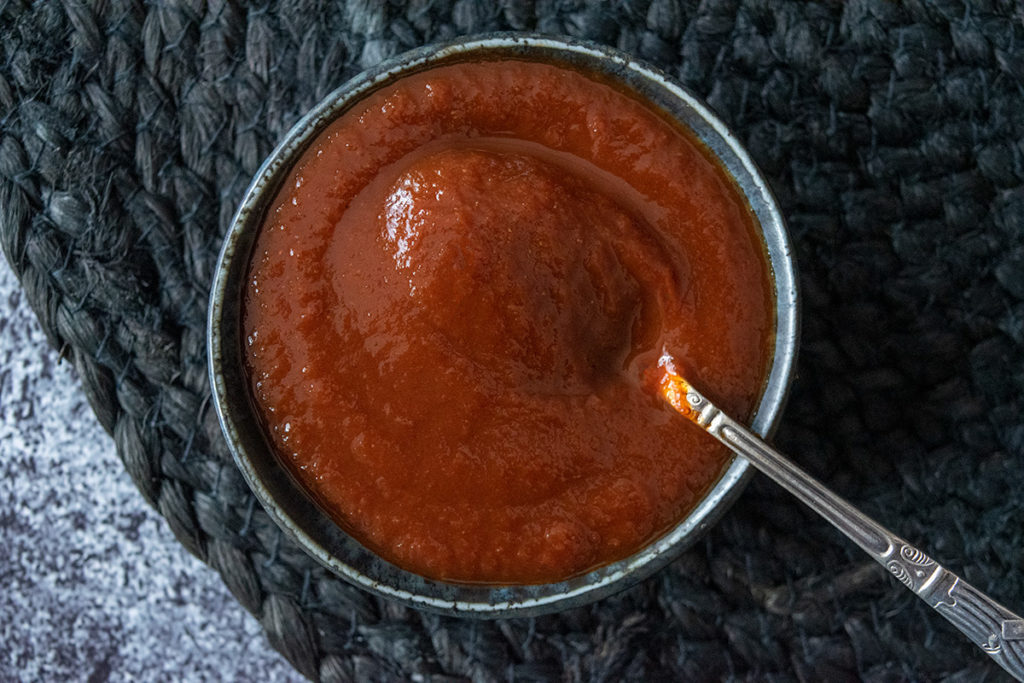 Thermomix bbq sauce in black ramekin on black textured round placemat, on rustic black tabletop. Spoon in dish, Birdseye view