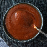 Thermomix bbq sauce in black ramekin on black textured round placemat, on rustic black tabletop. Spoon in dish, Birdseye view