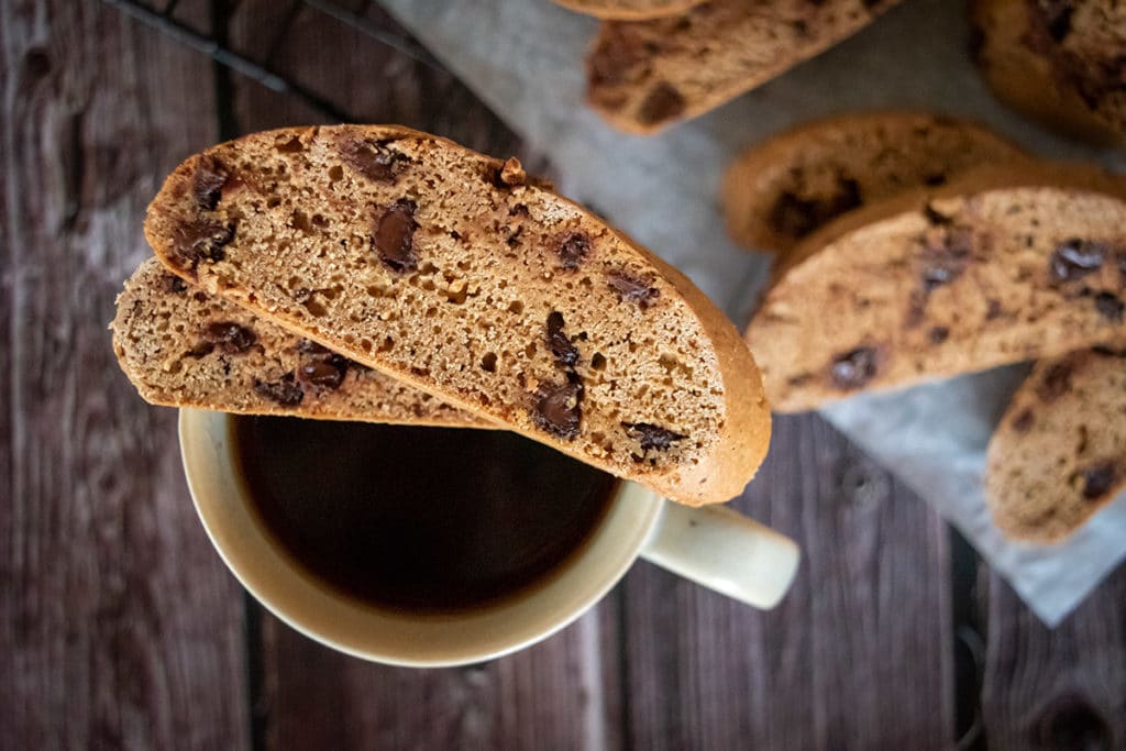 two slices choc chip biscotti made in Thermomix, sitting on top of white mug of black coffee. Sitting on wooden dark surface, with more biscotti in background, blurred.