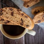 two slices choc chip biscotti made in Thermomix, sitting on top of white mug of black coffee. Sitting on wooden dark surface, with more biscotti in background, blurred.