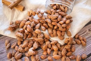 sugar and spice almonds spilling out of a jar, onto beige fabric on wooden surface, two cinnamon sticks on the left top