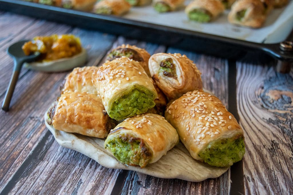 spinach and cheese rolls on rustic cream plate, on wooden table, ramekin of chutney with spoon and tray of more rolls in background