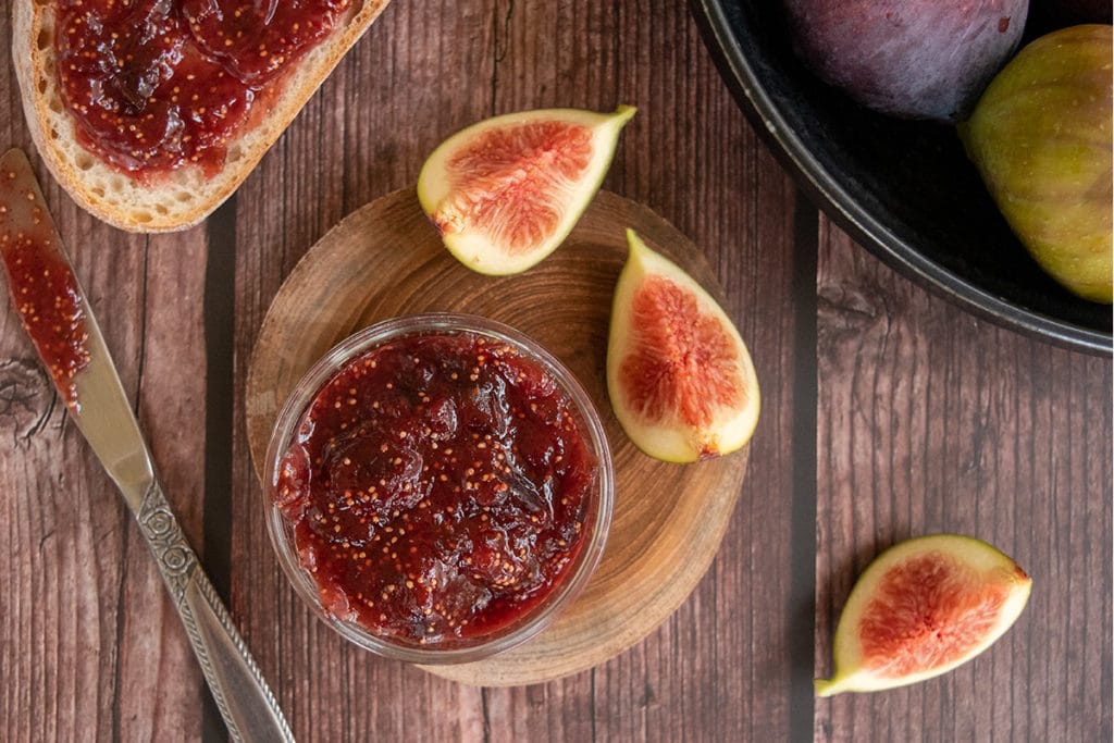 fig jam in small jar on wooden round plate, surrounded by sliced figs, whole figs and jam spread on bread. All on wooden table.