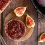 fig jam in small jar on wooden round plate, surrounded by sliced figs, whole figs and jam spread on bread. All on wooden table.