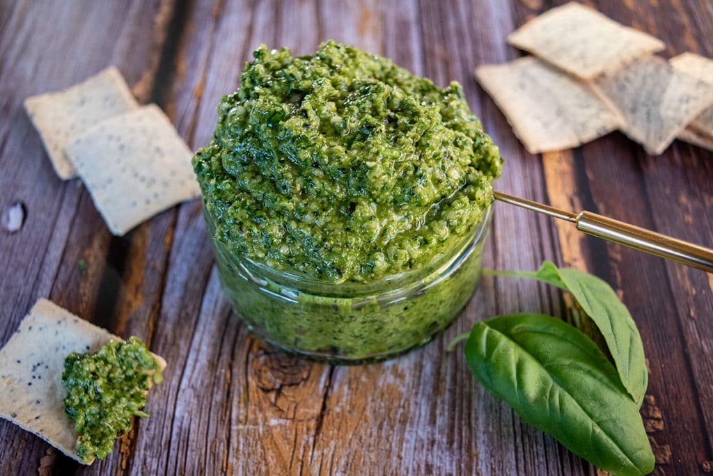 overflowing jar of hemp seed basil pesto, gold spoon handle sticking out, surrounded by crackers, two basil leaves in forefront