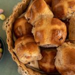 Basket packed full of hot cross buns that have been whipped up in thermomix, small bowl full of candied Easter eggs to the left with some on the tabletop also. Background is a green placemat and cream doily.