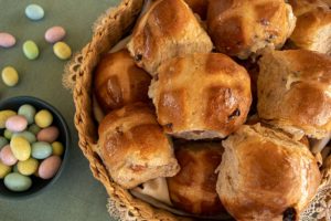 Basket packed full of hot cross buns that have been whipped up in thermomix, small bowl full of candied Easter eggs to the left with some on the tabletop also. Background is a green placemat and cream doily.