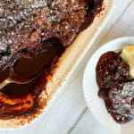 Birdseye view of chocolate self saucing pudding from thermomix in pudding dish, with spoon in sauce. Piece dished up on plate next to main dish, cream on portion. All on grey backdrop