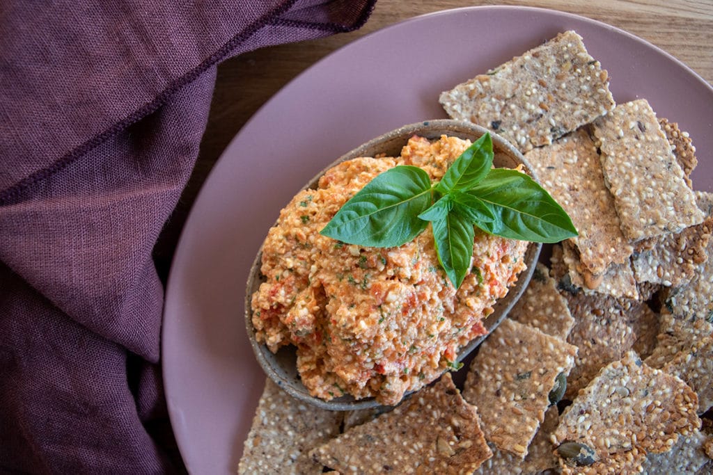 Pink plate with dish on top containing thermomix roasted capsicum and macadamia dip, with basil leaves on top, crackers around dip. Wooden tabletop with purple napkin to the left of the plate